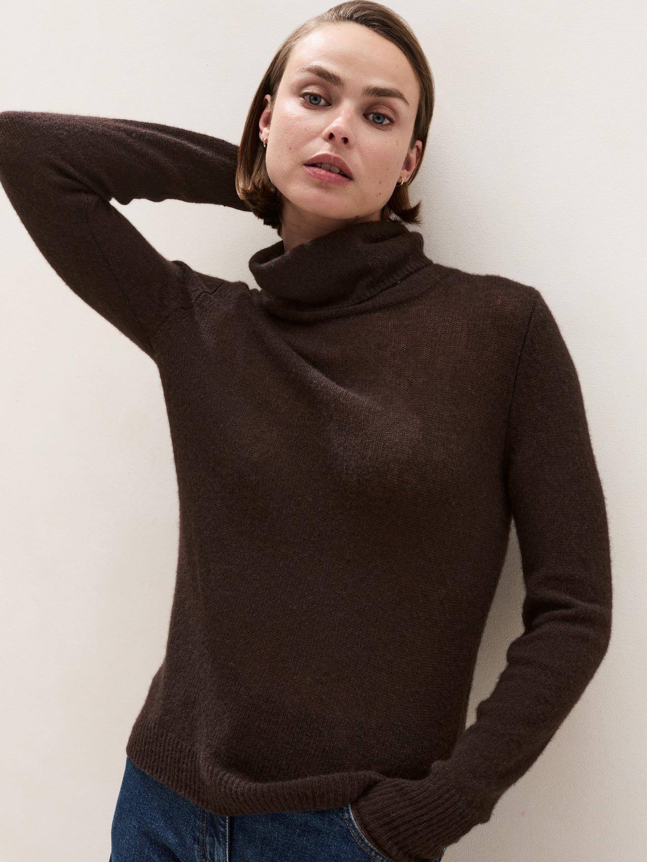 a woman in a brown turtle neck sweater leaning against a wall