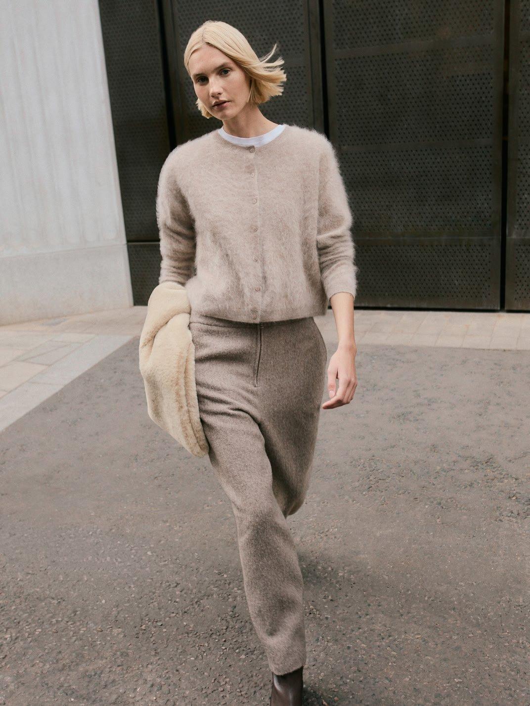 a woman walking down a street in a beige sweater and pants