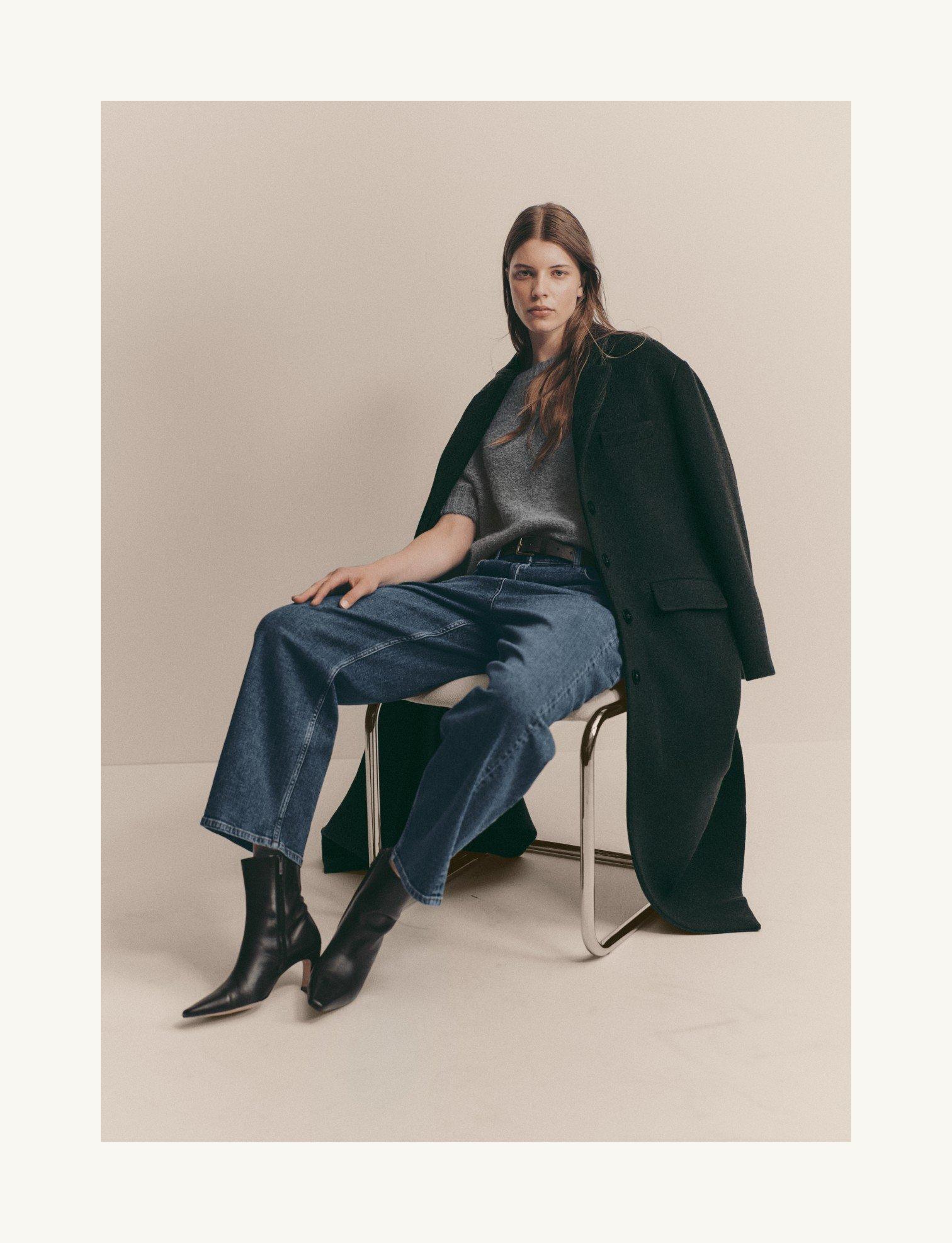 a woman sitting on a chair wearing jeans and a coat